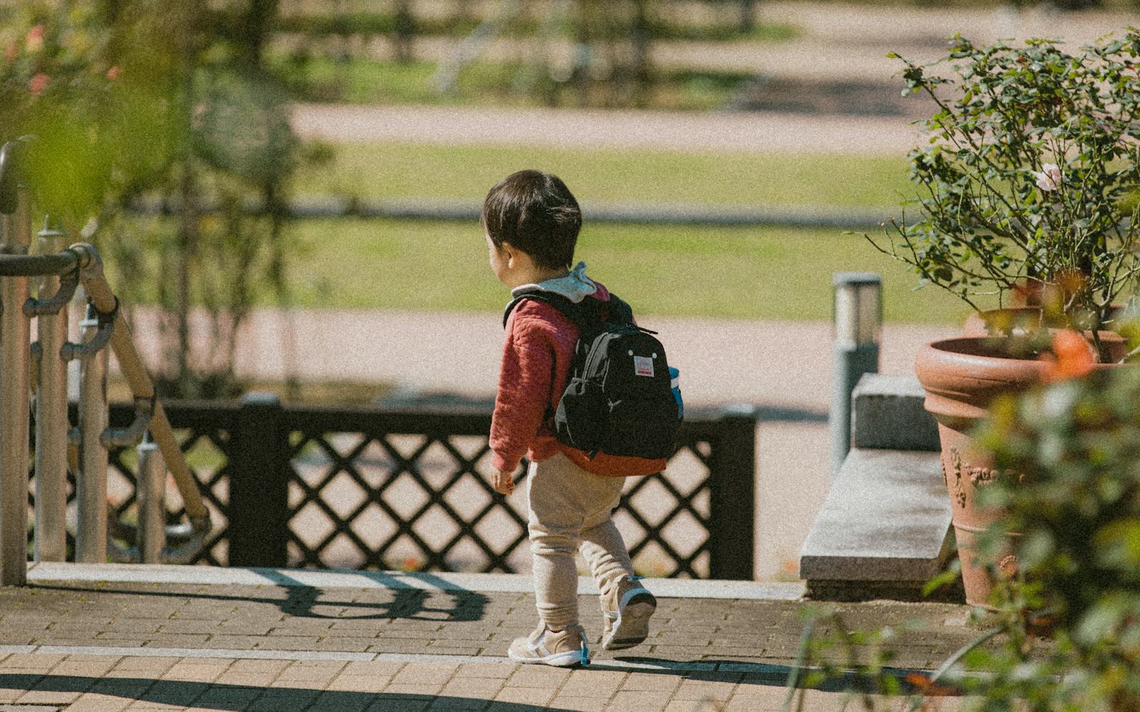 穿紅色外套、背著黑色小背包的幼兒，走在公園步道上的背影