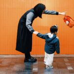橘色牆面前，媽媽牽著穿藍色連帽外套的幼兒，另一手提著橘色小背包