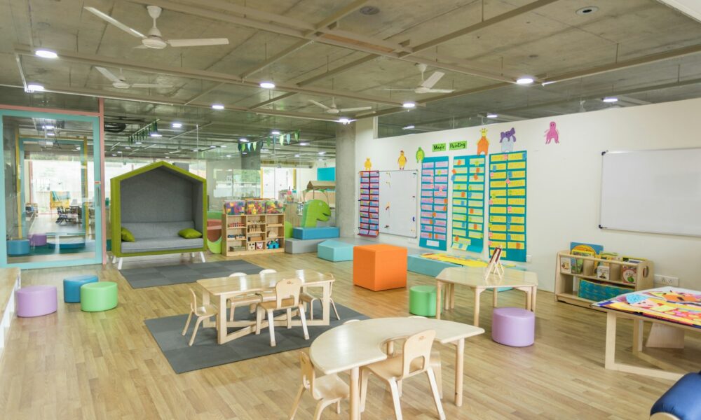 明亮的幼兒園教室，木地板上擺著低矮的桌椅、彩色坐墩與教具櫃，牆上貼著學習海報