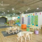 明亮的幼兒園教室，木地板上擺著低矮的桌椅、彩色坐墩與教具櫃，牆上貼著學習海報